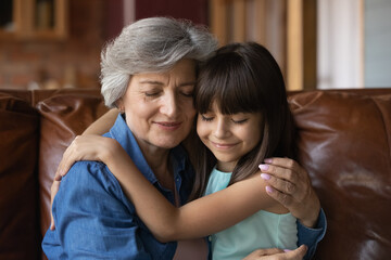 Close up mature woman and little girl hugging with closed eyes, sitting on couch at home, loving older grandmother and adorable granddaughter enjoying tender moment, embracing, two generations