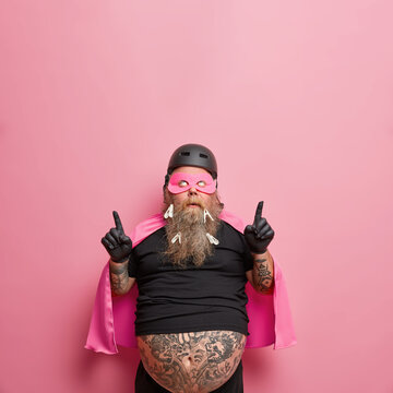 Overweight Tattooed Bearded Man With Big Belly Wears Superhero Costume Points Above On Copy Space Demonstrates Advertisement Ready To Organize Party For Children Thinks About Funny Entertainments