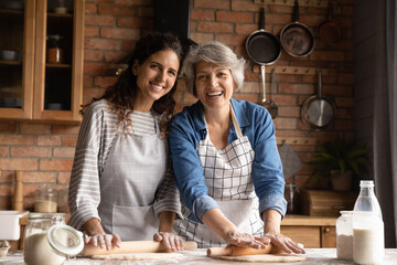 Head shot portrait smiling mature woman with grownup daughter wearing aprons cooking homemade pastry, standing in kitchen at home, happy family enjoying leisure time, preparing pie or bread