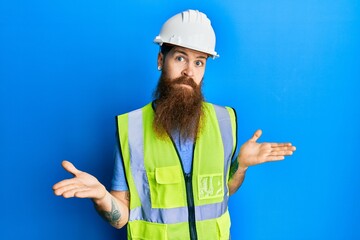 Redhead man with long beard wearing safety helmet and reflective jacket clueless and confused expression with arms and hands raised. doubt concept.