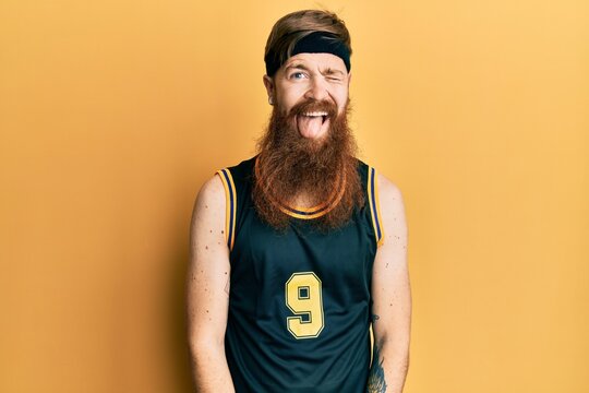 Redhead man with long beard wearing basketball uniform winking looking at the camera with sexy expression, cheerful and happy face.