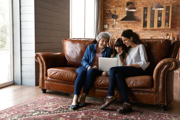 Smiling little girl with young mother and mature grandmother using laptop, sitting on couch at home, happy three generations of women enjoying leisure time, browsing apps, chatting or shopping online