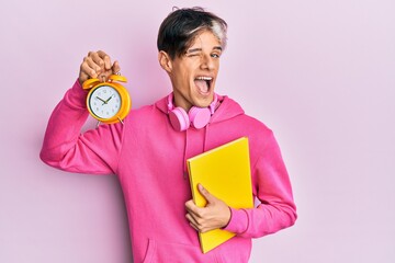 Young hispanic man holding book and alarm clock winking looking at the camera with sexy expression,...