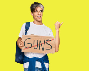 Young hispanic man holding no guns warning banner pointing thumb up to the side smiling happy with open mouth
