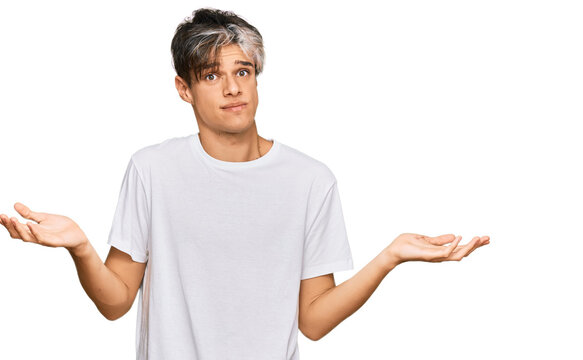 Young hispanic man wearing casual white tshirt clueless and confused expression with arms and hands raised. doubt concept.