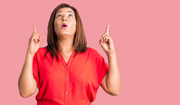 Middle age latin woman wearing casual clothes amazed and surprised looking up and pointing with fingers and raised arms.
