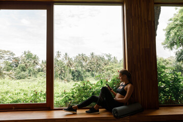 Young woman in sport clothes sit near the window with tropical view otdoor and palm trees, using mobile phone. Rolled yoga mat lie ear her, waiting for trainining
