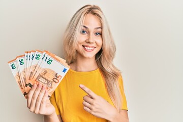 Beautiful caucasian blonde girl holding bunch of 50 euro banknotes smiling happy pointing with hand and finger - obrazy, fototapety, plakaty