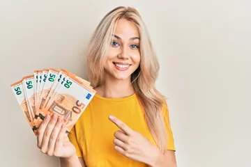 Fotobehang Beautiful caucasian blonde girl holding bunch of 50 euro banknotes smiling happy pointing with hand and finger © Krakenimages.com
