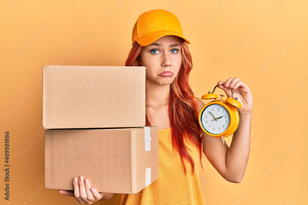 Poster young redhead woman holding delivery package and alarm clock depressed and worry for distress, cryin