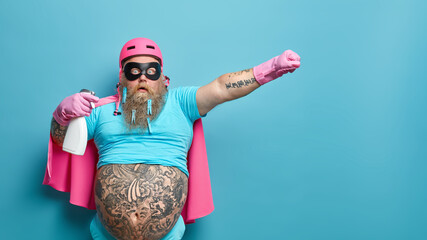 Startled cleaning superhero with detergent going to do washing up stares bugged eyes wears helmet face mask and cape has fat tattoed belly isolated over blue background. Time to tidy up room © WHstudio Leushin N