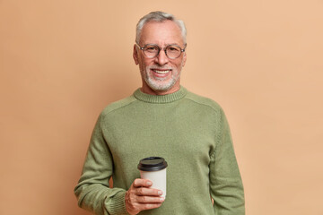 Horizontal shot of handsome bearded mature European man holds takeaway coffee enjoys pleasant conversation with interlocutor wears spectacles and casual sweater isolated over brown background