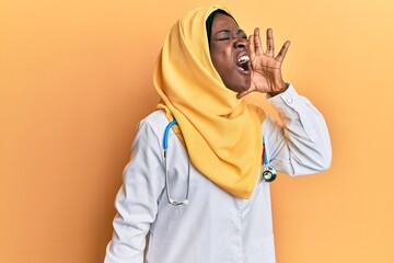 Beautiful african young woman wearing doctor uniform and hijab shouting and screaming loud to side with hand on mouth. communication concept.