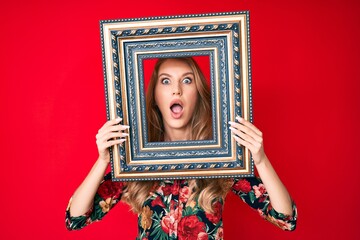 Young caucasian woman with blond hair holding empty frame afraid and shocked with surprise and amazed expression, fear and excited face.