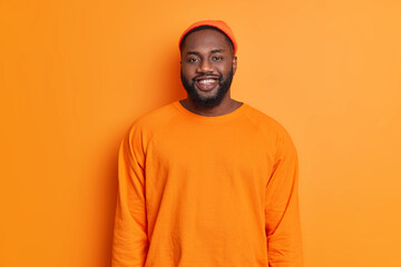Waist up shot of happy black man smiles happily dressed in orange hat and sweater being in good mood looks directly at camera expresses positive emotions stands in studio against bright background