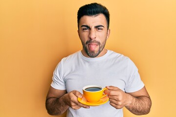 Young hispanic man drinking a cup of coffee sticking tongue out happy with funny expression.