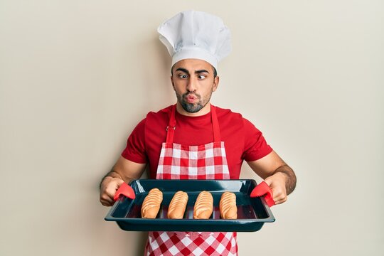 Young hispanic man wearing baker uniform holding homemade bread making fish face with mouth and squinting eyes, crazy and comical.