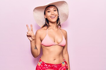 Young woman wearing bikini and hat showing and pointing up with fingers number two while smiling confident and happy.