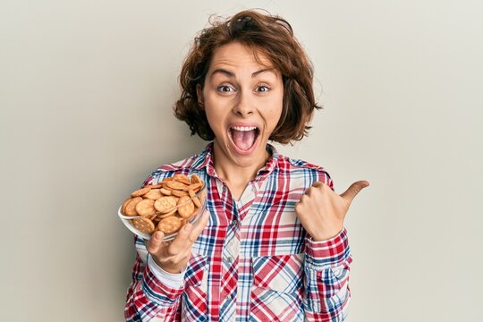Young brunette woman holding bowl with salty biscuits pointing thumb up to the side smiling happy with open mouth