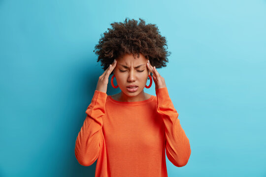 Photo of frustrated young Afro American woman has headache keeps hands on temples suffers unbearable migraine after noisy party wears orange sweater poses indoor isolated on blue background.