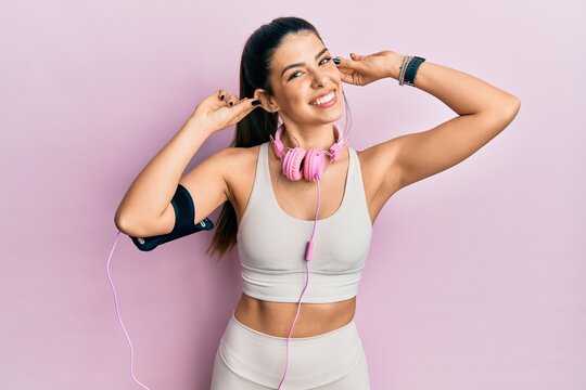 Young hispanic woman wearing gym clothes and using headphones smiling pulling ears with fingers, funny gesture. audition problem