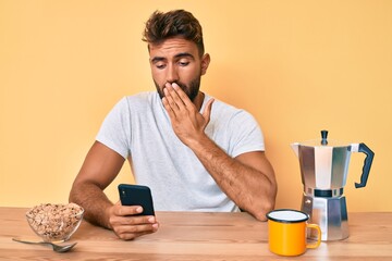 Young hispanic man sitting on the table having breakfast and using smartphone covering mouth with...