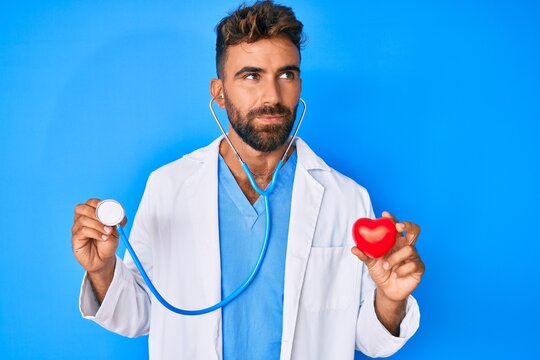 Young hispanic man wearing doctor uniform holding stethoscope and heart smiling looking to the side and staring away thinking.