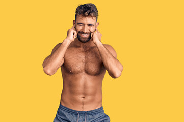Fototapeta premium Young hispanic man wearing swimwear shirtless covering ears with fingers with annoyed expression for the noise of loud music. deaf concept.