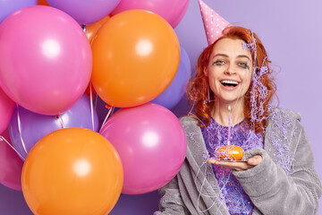 Fototapeta na wymiar Happy overjoyed redhead woman with leaked makeup enjoys pajama party at home wears birthday cone hat dressing gown smeared with cream smiles broadly holds inflated colorful balloons stands indoor
