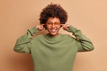 Fototapeta na wymiar Positive dark skinned curly haired woman smiles happily plugs ears avoids very loud music wears transparent glasses and sweater has happy mood isolated over brown background. Its too noisy here