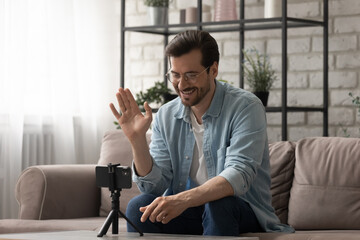 Fototapeta premium Smiling man wearing glasses waving hand, using smartphone on tripod, popular blogger recording vlog, sitting on couch at home, greeting followers, shooting video for channel in social network