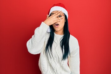 Beautiful hispanic woman wearing christmas hat peeking in shock covering face and eyes with hand, looking through fingers afraid