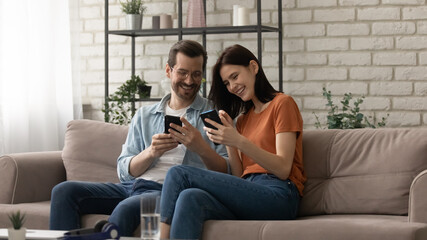 Overjoyed couple using phones together, sitting on cozy couch at home, happy man wearing glasses and woman surfing internet, holding smartphones, having fun with gadgets, enjoying leisure time