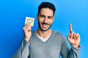 Young hispanic man holding heart reminder smiling with an idea or question pointing finger with...