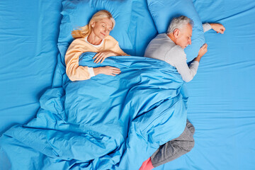 Sweet dreams and napping concept. Sleeping aged couple pose in bed under blanket fall asleep at bedroom enjoys serene atmosphere dressed in nightwear. Senior woman and man have rest at night