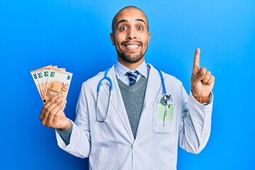 Hispanic adult doctor man wearing medical uniform holding 50 euros banknotes smiling with an idea or question pointing finger with happy face, number one