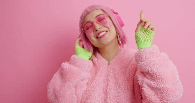 Carefree pink haired Asian woman wears stereo headphones dressed in winter coat and sport gloves dances with rhythm of music isolated over pink background. People happiness fun hobby concept