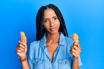Beautiful hispanic woman eating chicken wings in shock face, looking skeptical and sarcastic,...