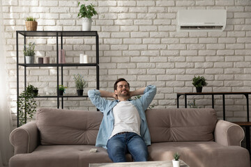 Calm man wearing glasses relaxing with closed eyes in room with air conditioner, sitting leaning back on cozy couch at home, breathing fresh air, satisfied customer enjoying comfortable temperature - obrazy, fototapety, plakaty