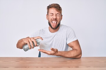 Young caucasian man using hand sanitizer gel sitting on the table sticking tongue out happy with funny expression.