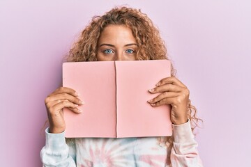Beautiful caucasian teenager girl reading a book covering face sticking tongue out happy with funny expression.