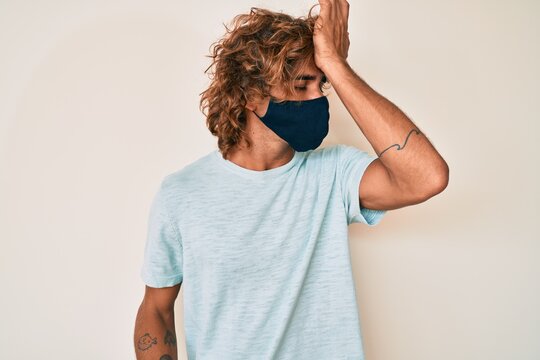 Young hispanic man wearing covid-19 mask surprised with hand on head for mistake, remember error. forgot, bad memory concept.