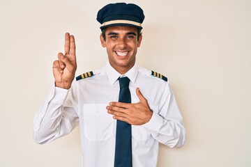 Young hispanic man wearing airplane pilot uniform smiling swearing with hand on chest and fingers up, making a loyalty promise oath