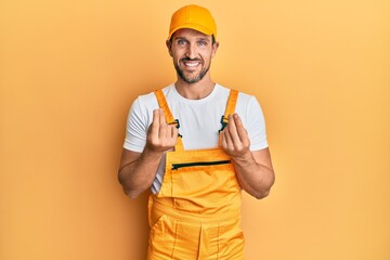 Young handsome man wearing handyman uniform over yellow background doing money gesture with hands,...