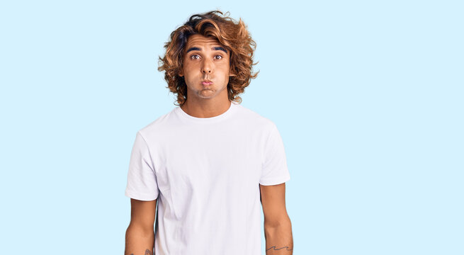 Young hispanic man wearing casual white tshirt puffing cheeks with funny face. mouth inflated with air, crazy expression.