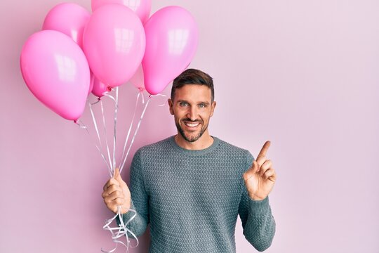 Handsome man with beard holding pink balloons smiling happy pointing with hand and finger to the side