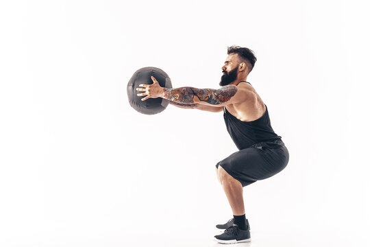 Muscular tattooed bearded male exercising fitness weights Medicine Ball in studio isolated on white background.