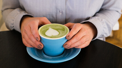 Woman holding blue cup with hot trendy green latte with art heart on the foam on black wooden table background, healthy drink with latte art