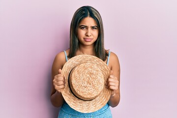 Young latin woman holding summer hat clueless and confused expression. doubt concept.
