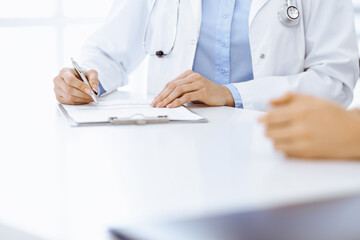 Unknown woman-doctor and female patient sitting and talking at medical examination in clinic, close-up. Therapist wearing blue blouse is filling up medication history record. Medicine concept